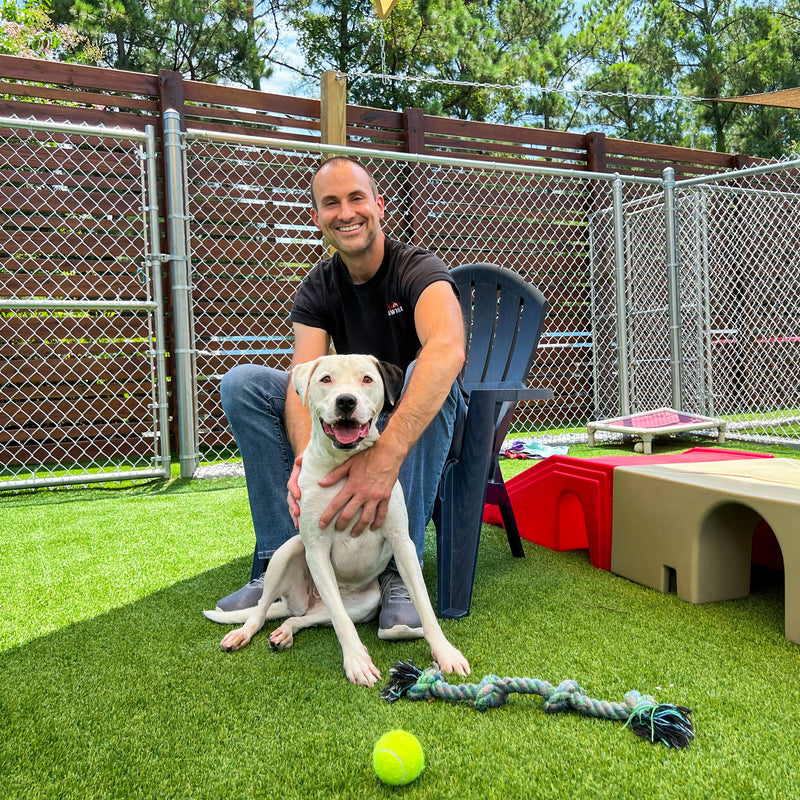 Man sitting with a dog in an outdoor play area with toys and a fence in the background